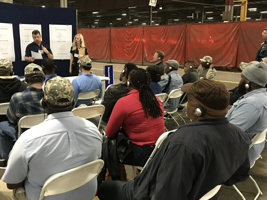Seated audience wearing headsets, listening to presenters in a plant