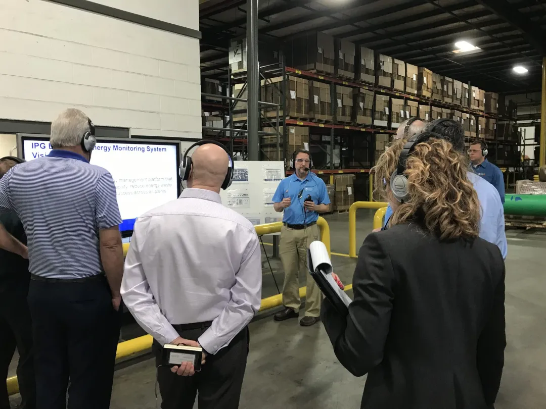 A presenter with a microphone speaks to a tour group wearing headsets in a warehouse