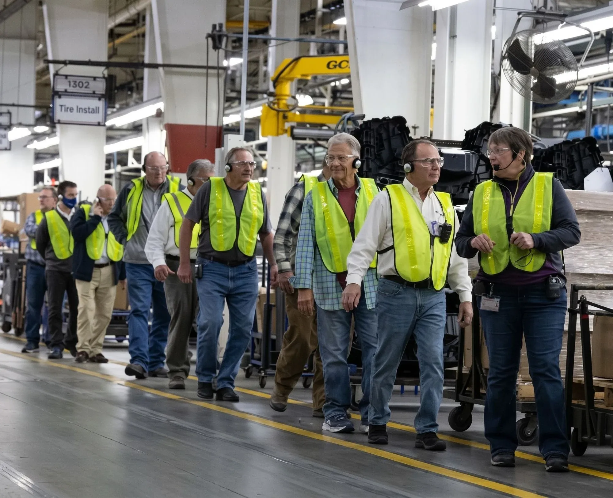 Tour group in safety vests and glasses wearing headsets, following a guide with a headset microphone along a factory floor