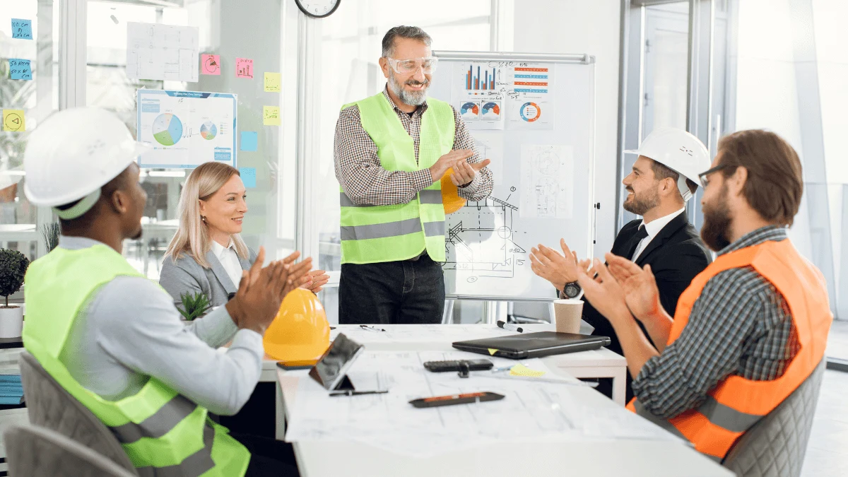 Workers in hard hats and hi-vis vests applauding a colleague during a meeting around a conference table