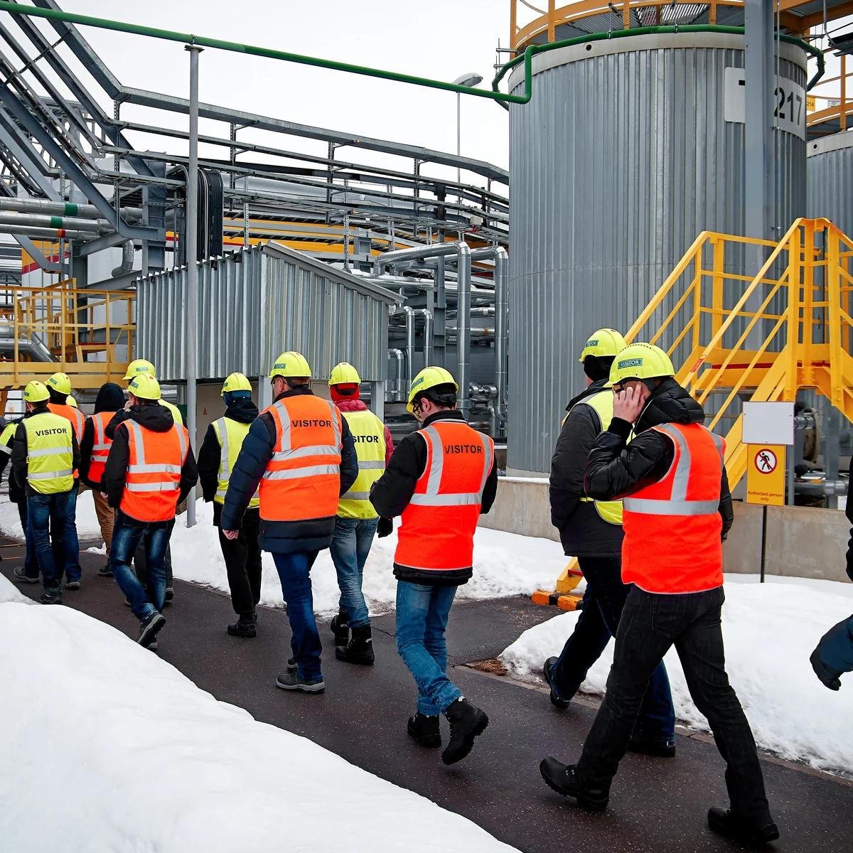 Group of visitors in hard hats and high-visibility vests walking through an industrial plant in snow
