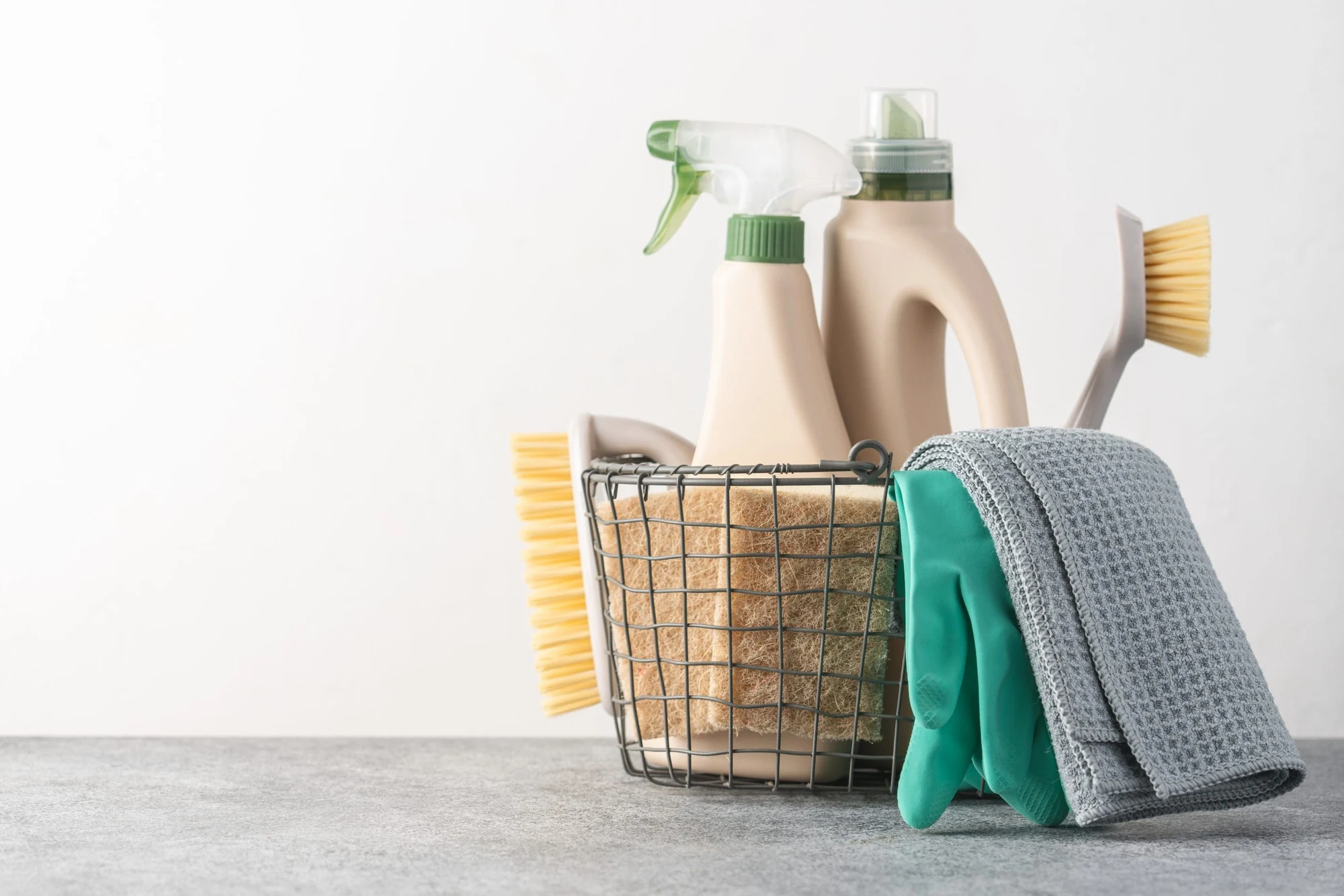 Wire basket of cleaning supplies with spray bottles, scrub brush and cloths on a table