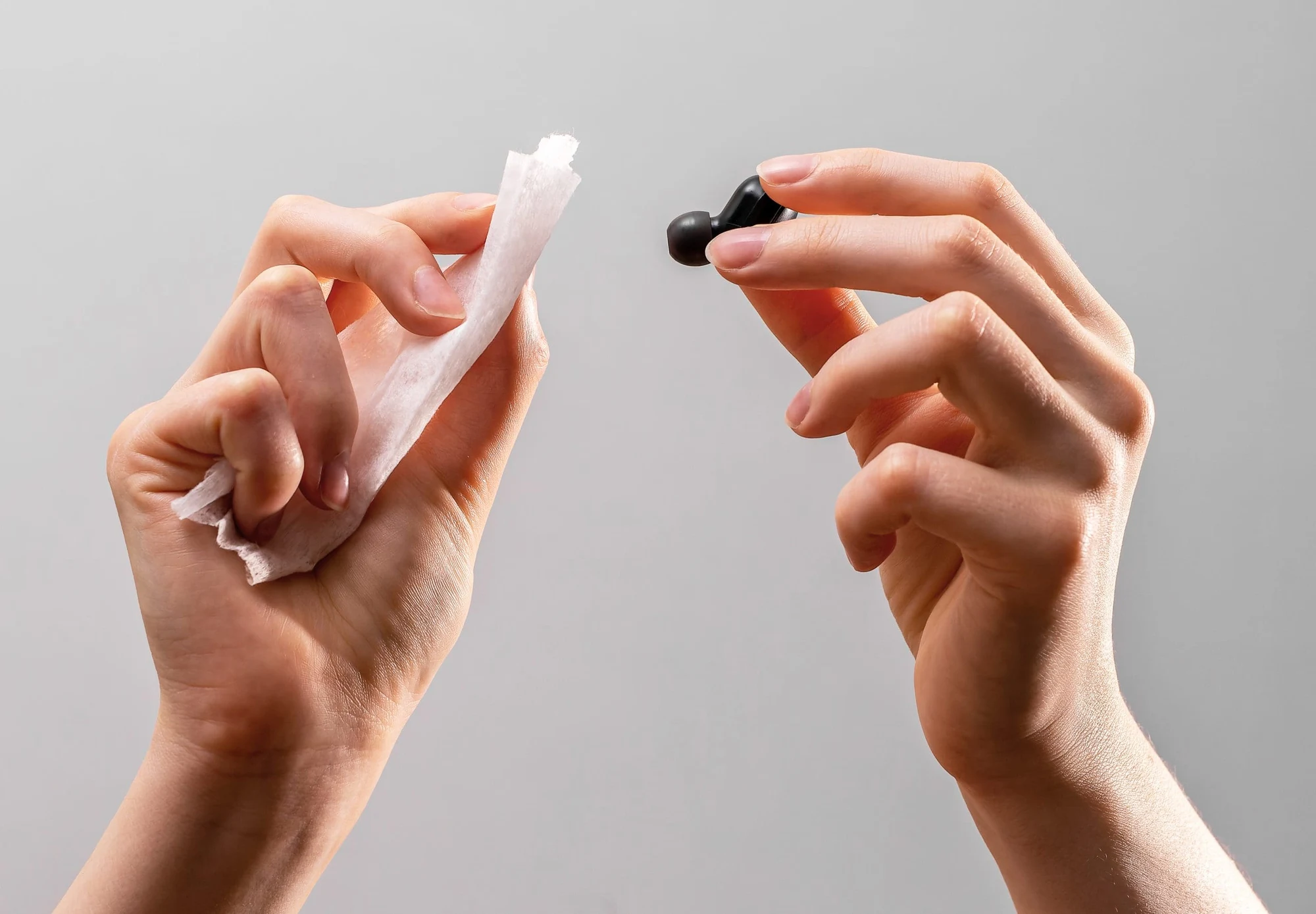 Hands cleaning a small black earbud with a white cloth