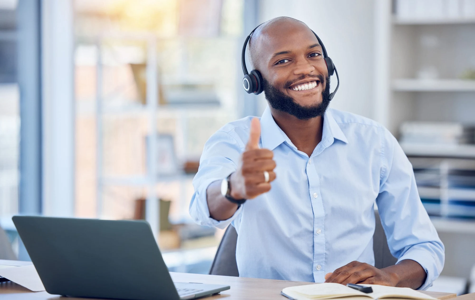 Smiling man wearing a headset giving a thumbs up at a desk with a laptop