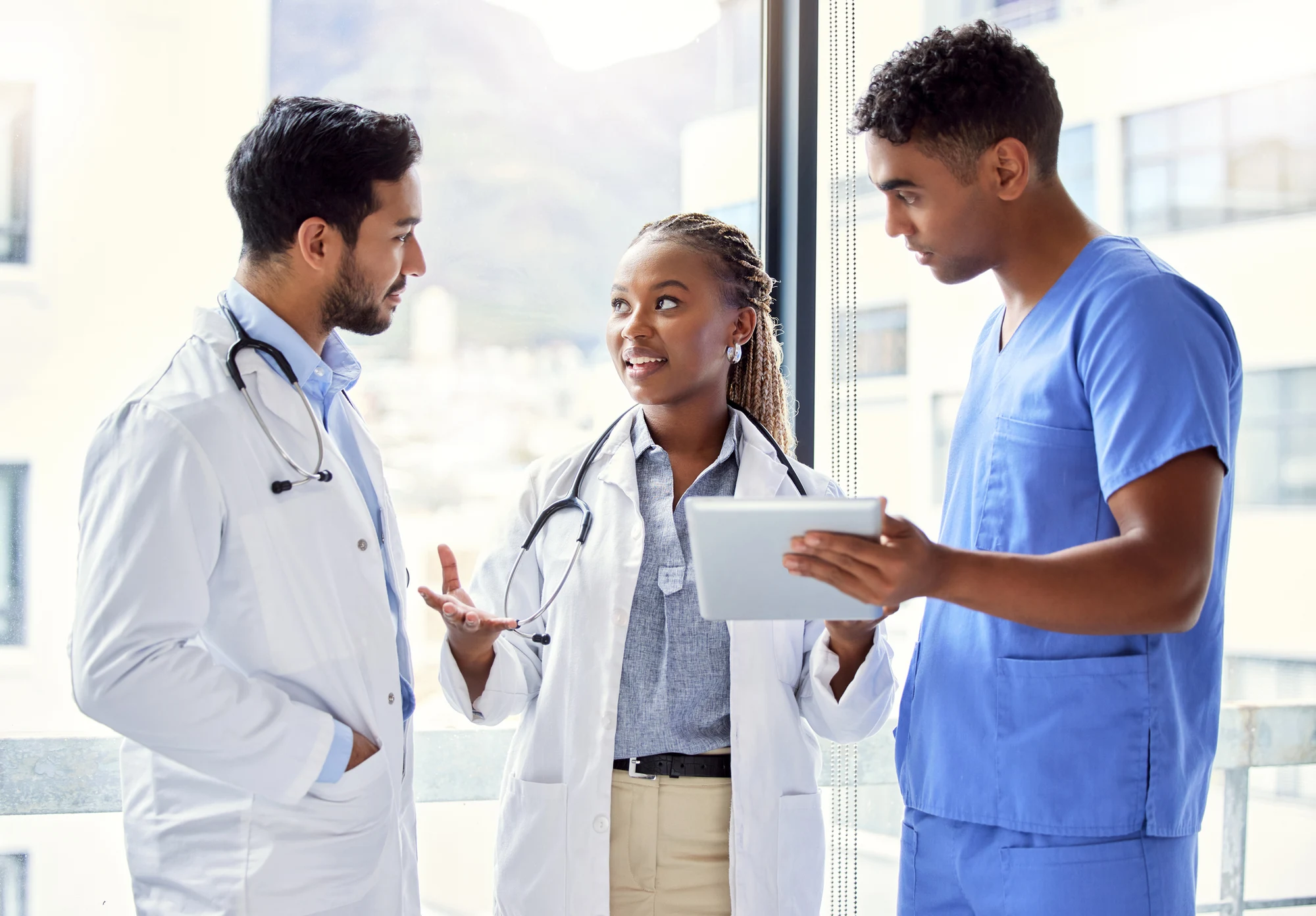 Two doctors in white coats and a nurse in blue scrubs talking while one holds a tablet