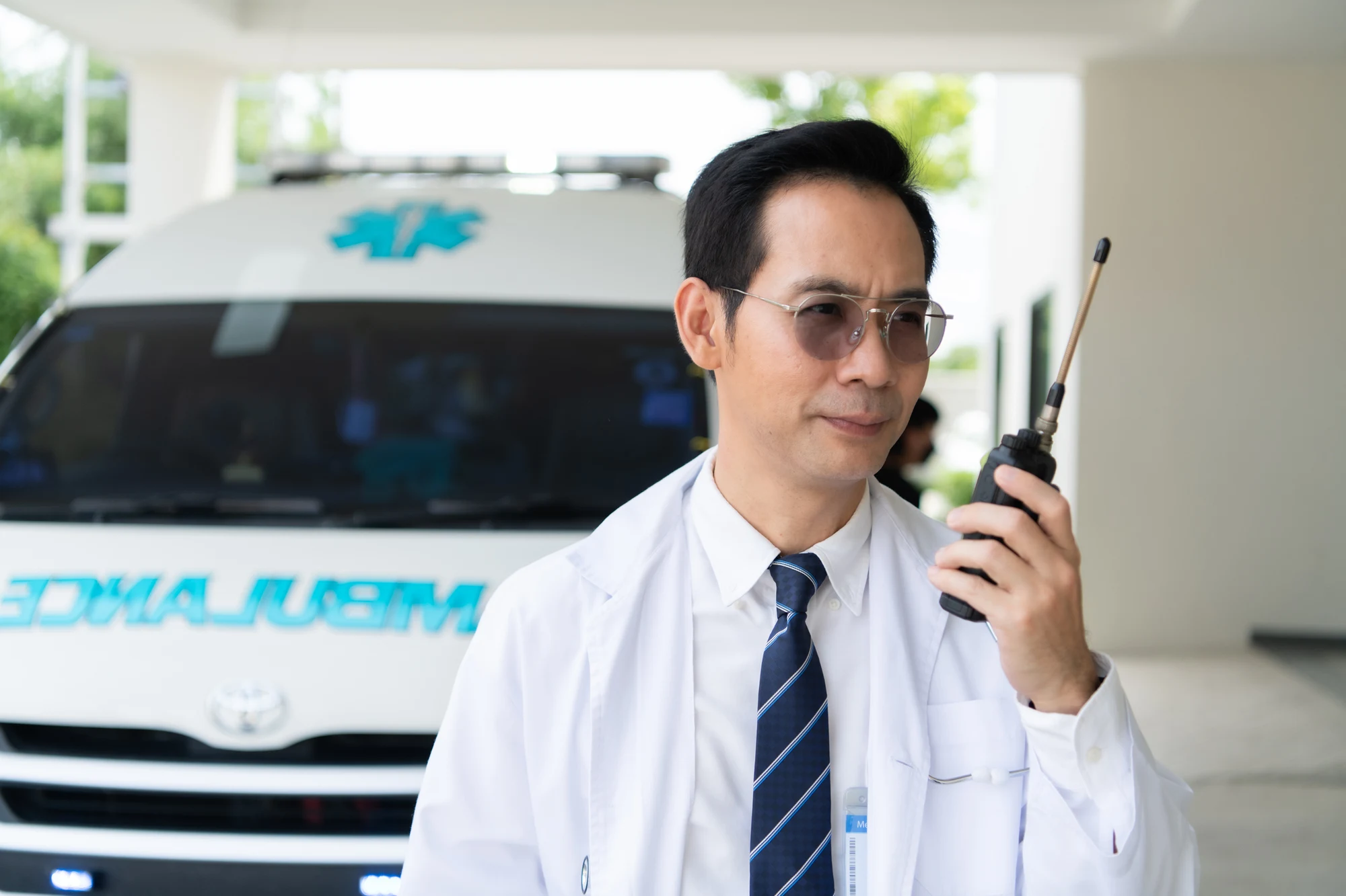 Man in a white coat and tie speaking into a two-way radio in front of an ambulance