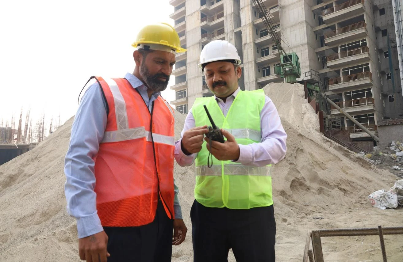 Two workers in hard hats and safety vests looking at a two-way radio at a construction site