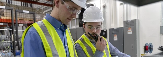Two workers in hard hats, safety glasses and hi-vis vests, one speaking into a two-way radio