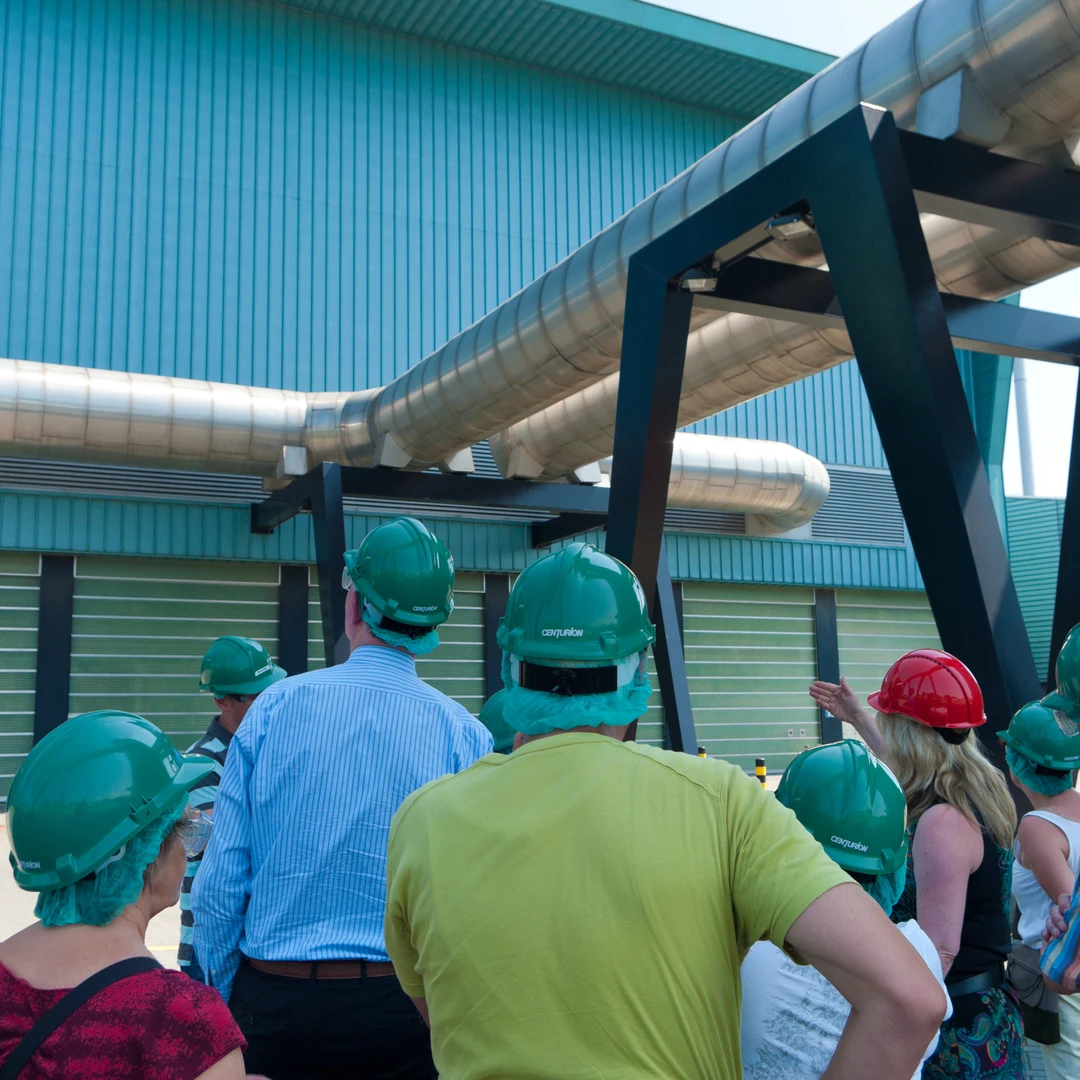 Tour group in green hard hats outside an industrial building with overhead pipework