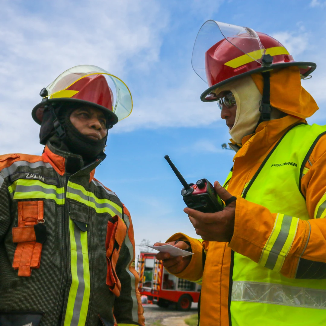 Two firefighters in helmets and gear, one using a handheld two-way radio